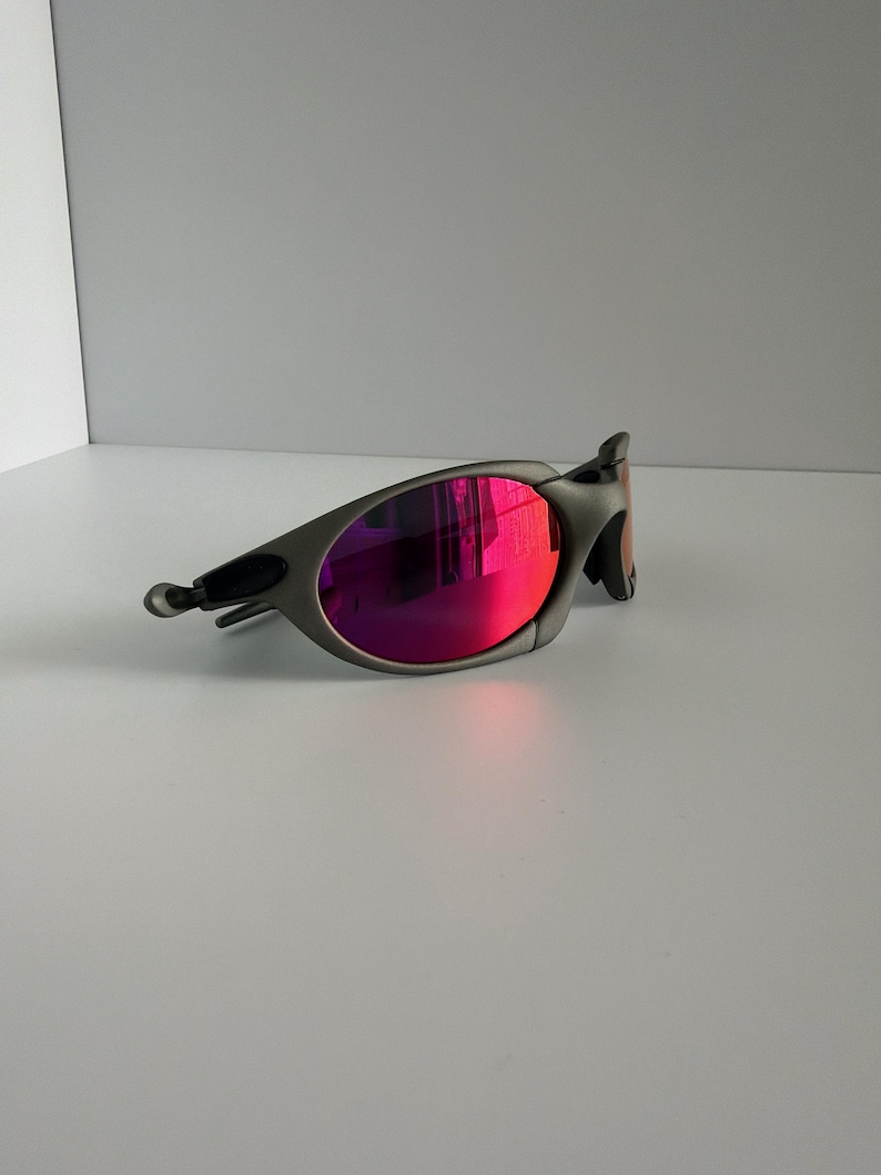 May include: A pair of gray sunglasses with vibrant pink lenses. The sunglasses have a sleek, modern design with a curved frame and a small, curved arm. The lenses reflect a bright pink hue, adding a pop of color to the overall look.