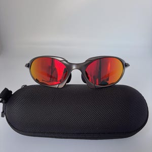 May include: Dark grey sunglasses with red mirrored lenses sit on a black, oval case. The sunglasses have a curved frame and a modern design. The case has a textured surface and a zip closure.