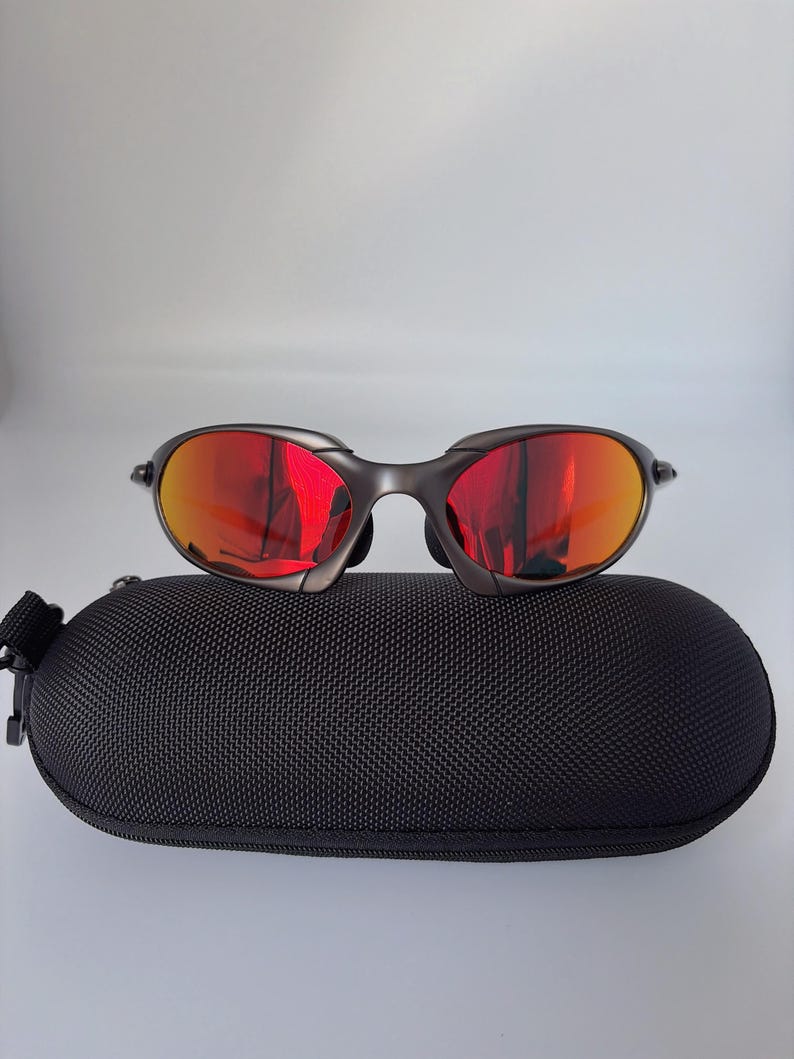 May include: Dark grey sunglasses with red mirrored lenses sit on a black, oval case. The sunglasses have a curved frame and a modern design. The case has a textured surface and a zip closure.