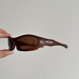May include: Brown sunglasses with a curved frame and dark lenses. The sunglasses have a matte finish and a silver logo on the temple. The design is sporty and modern.