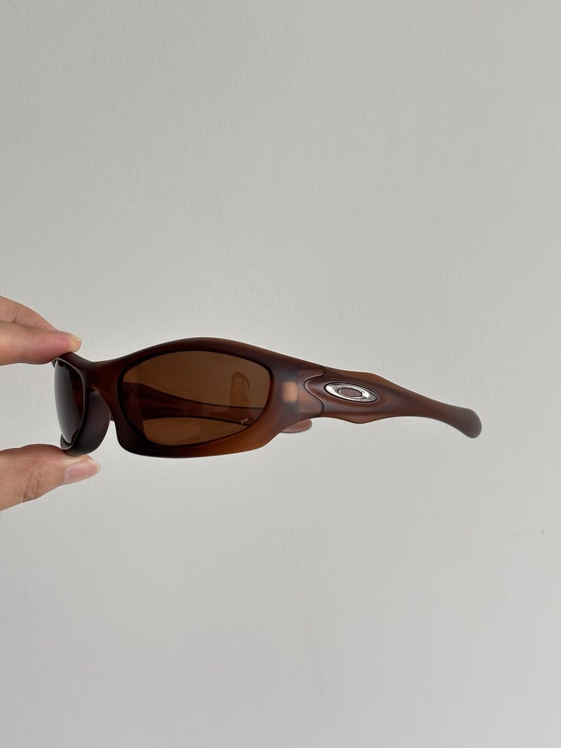 May include: Brown sunglasses with a curved frame and dark lenses. The sunglasses have a matte finish and a silver logo on the temple. The design is sporty and modern.