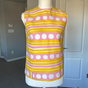 May include: A sleeveless top with a vibrant pattern of horizontal yellow and pink stripes, interspersed with rows of large white polka dots. The top has a round neckline and small side slits. The fabric appears to be a lightweight material.