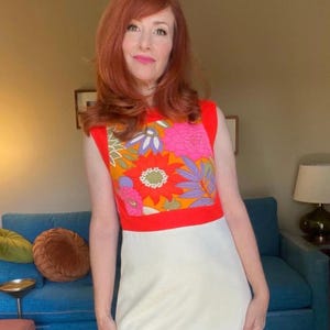 Może przedstawiać: Sukienka bez rękawów z odważnym kwiatowym nadrukiem na gorsecie. Góra jest w kolorze jasnoczerwonym z kwadratowym kwiatowym wzorem w kolorze pomarańczowym, różowym i fioletowym. Spódnica jest w jednolitym kremowym kolorze, tworząc uderzający kontrast. Sukienka ma estetykę retro i vintage.