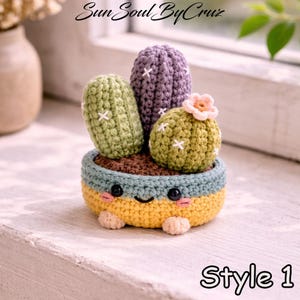 May include: Crocheted cactus plant in a smiling pot. The cactus features three different shapes in green and purple, with white cross stitches. The pot is yellow, blue, and brown, with a smiling face and the text "Style 1".