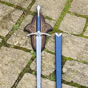 Op de afbeelding: Een decoratief zwaard met een zilveren lemmet en een blauw handvat, gemonteerd op een houten plaat. De plaat heeft een donkerbruine afwerking met goudkleurige decoratieve accenten. Een blauwe schede staat naast het zwaard.