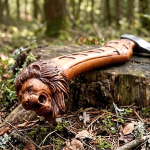 May include: A handcrafted axe with a wooden handle featuring a detailed lion's head carving. The axe head is silver, and the handle is a warm brown color. The axe is resting on a tree stump in a forest setting.