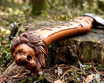 May include: A handcrafted axe with a wooden handle featuring a detailed lion's head carving. The axe head is silver, and the handle is a warm brown color. The axe is resting on a tree stump in a forest setting.