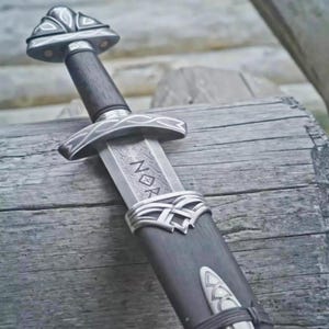 Peut inclure: Épée viking avec une poignée en bois sombre et une garde argentée. La lame présente des gravures, dont les lettres "NOR". L'épée est gainée dans un fourreau sombre, posée sur une surface en bois, avec une esthétique rustique.