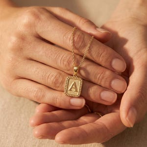 Könnte beinhalten: Eine goldfarbene Halskette mit einem Anhänger mit dem Buchstaben "A" in einem quadratischen Rahmen. Der Anhänger hängt an einer zarten Kette. Die Halskette wird in offenen Händen gehalten.