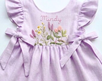 May include: A lavender-colored dress with embroidered floral details and the name "Mindy". The dress features ruffled sleeves and tie closures at the waist. The embroidery includes daisies, lavender, and pink flowers.