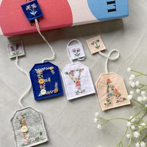Può includere: Etichette ricamate per bustine di tè in vari colori, tra cui blu, bianco e grigio. Ogni etichetta presenta una lettera dell'alfabeto, decorata con ricami floreali colorati. Le etichette sono collegate da fili bianchi.