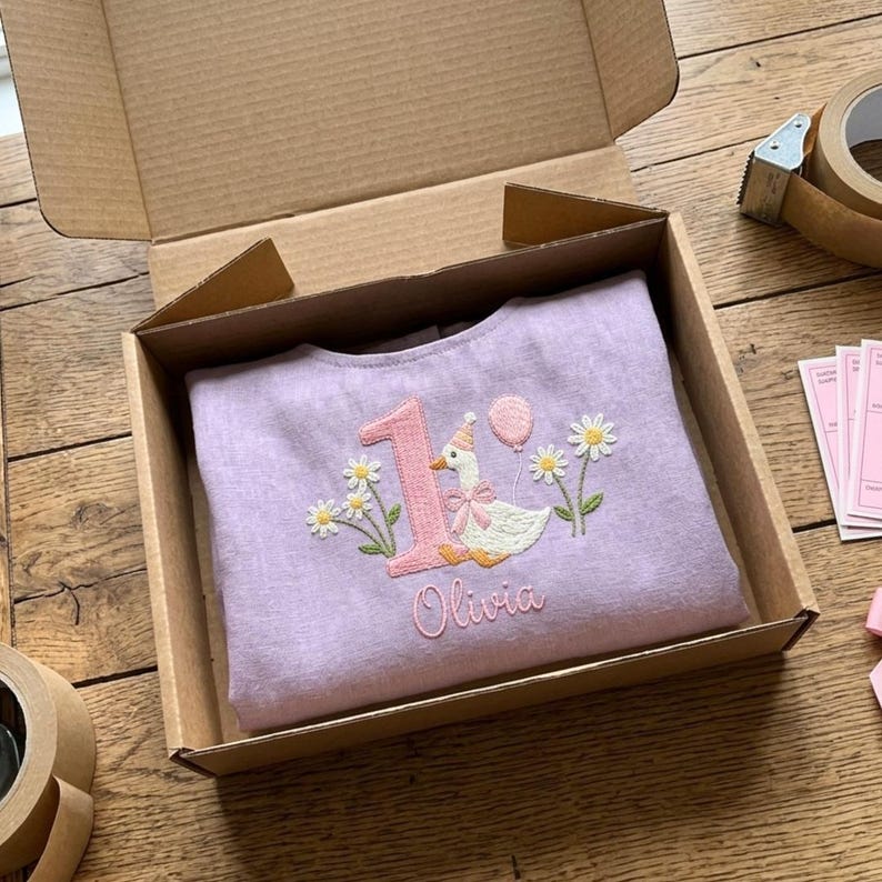 May include: A lilac-colored baby shirt with an embroidered design featuring a number 1, a goose wearing a party hat and bow, a balloon, and the name "Olivia." The shirt is neatly folded inside a cardboard box.