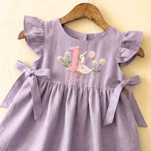 May include: A lavender linen dress with ruffled sleeves and bow ties. The dress features an embroidered design of a goose wearing a party hat, the number 1, daisies, a balloon, and the name "Olivia".