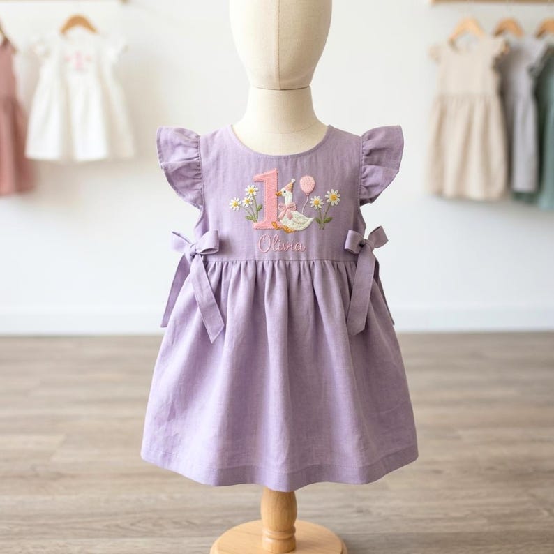 May include: A lavender dress with flutter sleeves and bow accents. The dress features an embroidered design with the number "1", a goose, daisies, and the name "Olivia". Perfect for a first birthday celebration.