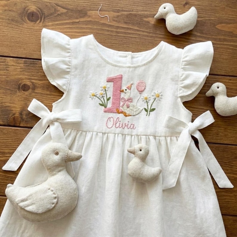 May include: White linen dress with ruffle sleeves and bow ties. The dress features an embroidered design with the number "1", a goose, daisies, and the name "Olivia" in pink thread. Includes felt duck decorations.
