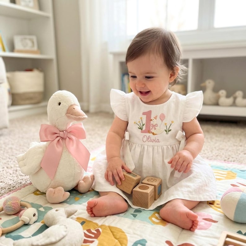 May include: A white dress with a pink number 1 and the name "Olivia" embroidered on it. A plush white duck with a pink bow sits next to the child. The child is playing with wooden blocks on a colorful mat.