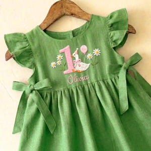 May include: Green linen dress with ruffled sleeves and bow ties. The dress features an embroidered design of a goose wearing a party hat, the number 1, daisies, a balloon, and the name "Olivia".