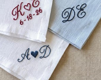 May include: Three embroidered handkerchiefs in white and light blue. The white handkerchiefs feature burgundy and navy blue embroidered initials and dates. The light blue handkerchief has navy blue embroidered initials. The embroidery includes hearts and dates.