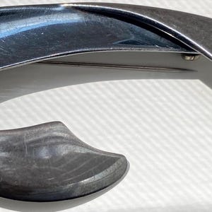 May include: A dark gray metallic brooch featuring a curved, abstract crescent shape with a smooth, polished finish. A small, spherical bead is attached to the upper left section of the frame, and a thin metal pin is visible on the back.