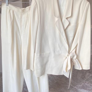 May include: A cream-colored two-piece suit consisting of a wrap-style blazer and matching wide-leg trousers. The blazer features long sleeves, a notched lapel, a single chest pocket, and a side tie closure. The trousers have a high waist.