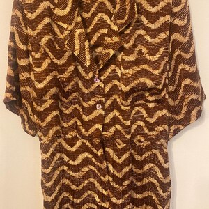 May include: A short-sleeved button-down shirt featuring a dark brown and beige wavy zigzag pattern. The garment has a notched collar, two front buttons, and a visible brand label at the inner neckline. The fabric displays a consistent repeating print.