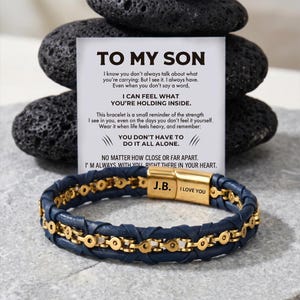 May include: A navy blue braided leather bracelet with a gold-tone metal chain centre and a gold-tone clasp engraved with J.B. and I LOVE YOU. It is displayed with a white card featuring the text TO MY SON and a message about strength and support.