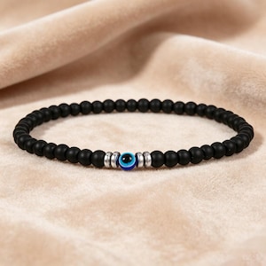 May include: Black beaded bracelet with a central blue and white evil eye charm. The bracelet features small, round, matte black beads. The evil eye charm is surrounded by silver-colored metal spacers.