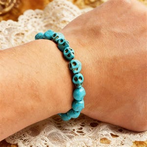 Könnte beinhalten: Ein türkisfarbenes Schädelperlenarmband. Das Armband besteht aus mehreren schädelartigen Perlen, die aneinandergereiht sind. Die Perlen haben eine leuchtende türkisfarbene Farbe, und die Schädel haben detaillierte Gesichtszüge. Das Armband wird am Handgelenk getragen.