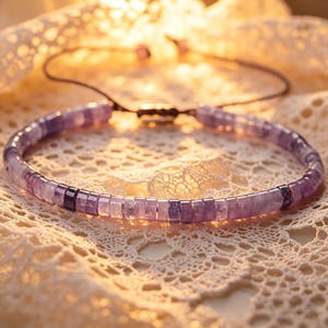 May include: A delicate bracelet featuring small, square-cut amethyst beads in varying shades of purple. The bracelet is strung on a dark cord with an adjustable knot closure. The background is a soft, cream-colored lace.