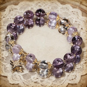 May include: A beaded bracelet featuring alternating purple and clear crystal beads. The bracelet has gold-colored accents and a star-shaped charm. The beads are round and vary in shades of purple.