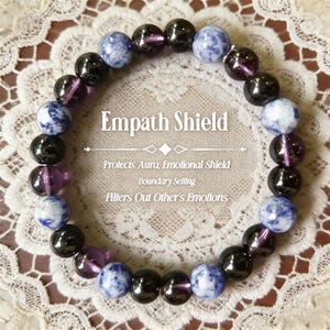 May include: A bracelet with the text "Empath Shield". The bracelet is made of black, purple, and blue-grey spherical beads. The text below the title reads "Protects Aura, Emotional Shield, Boundary Setting, Filters Out Other's Emotions."