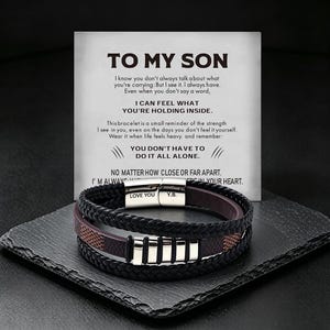 May include: A multi-strand black braided leather bracelet with a silver-tone magnetic clasp engraved with LOVE YOU and Y.B. The bracelet rests on a card with the text TO MY SON, I know you don't always talk about what you're carrying, and more.