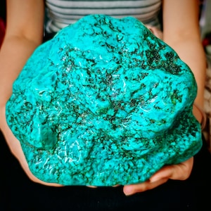 May include: A large, raw turquoise stone featuring a vibrant, bright blue-green color with intricate, dark black matrix webbing throughout its uneven, textured surface. The mineral specimen displays a natural, organic shape with varied contours.