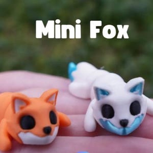 Puede incluir: Dos pequeñas figuras de zorro, una naranja y blanca y otra blanca y azul, con ojos y narices negras. El texto "Mini Fox" aparece en letras blancas sobre las figuras. Las figuras están esculpidas con orejas puntiagudas y posición tumbada.