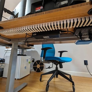 May include: A light-colored wooden cable management tray mounted beneath a bamboo desk surface. The tray features a series of vertical slats for organizing cords and power cables, with a black power strip and electronic components attached nearby.