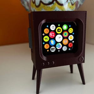 May include: A dark brown, 3D-printed stand shaped like a vintage television set, designed to hold a smartwatch. The stand features four tapered legs, a rectangular screen opening, and three circular buttons on the front panel below the display.