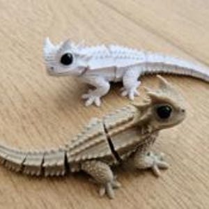 Puede incluir: Dos figuras de lagarto articuladas con grandes ojos negros y crestas dorsales espinosas. Un lagarto es de color blanco sólido y el otro es de color tostado claro. Ambas figuras cuentan con colas y cuerpos segmentados que permiten el movimiento.
