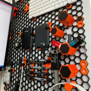 Op de afbeelding: Een zwart zeshoekig wandrooster met oranje montageclips en hulpstukken. Het systeem houdt diverse voorwerpen vast, waaronder externe harde schijven, USB-sticks, pennen, een digitale schuifmaat en een wit toetsenbord, wat een modulaire opbergoplossing vormt.