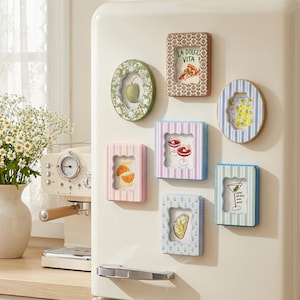 Può includere: Una collezione di sei calamite a forma di cornice rettangolare e ovale attaccate a un frigorifero color crema. Le cornici presentano vari motivi, tra cui righe e fiori, con illustrazioni di cibi e bevande e il testo LA DOLCE VITA.