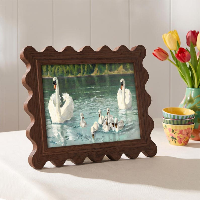 May include: A framed print featuring a serene lake scene with swans and cygnets. The dark brown frame has a wavy, scalloped edge. A vase of tulips and decorative bowls are in the background, adding a touch of color to the composition.