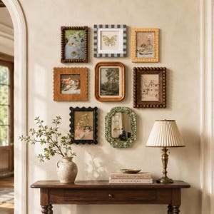 May include: A gallery wall arrangement featuring eight assorted rectangular picture frames with varied textures and colors, including beaded, scalloped, and checkered designs, surrounding a central landscape print, above a wooden console table.