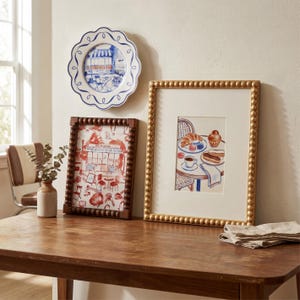 May include: A decorative arrangement featuring a blue and white plate with a cafe scene, two framed artworks, and a small vase with greenery. One frame has a beaded border, the other a gold beaded border. The artworks depict cafe scenes.