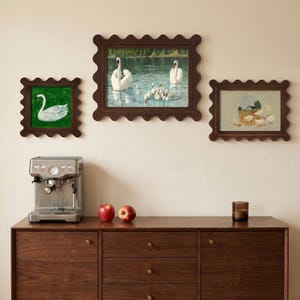 May include: Three framed artworks with scalloped edges depict swans and a farm scene. A stainless steel espresso machine, two red apples, and a small brown glass sit on a dark wood cabinet.