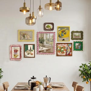 May include: A gallery wall arrangement featuring a central scalloped pink mirror surrounded by various rectangular frames containing art prints of food, plants, and still life scenes, illuminated by a cluster of five gold pendant lights above.