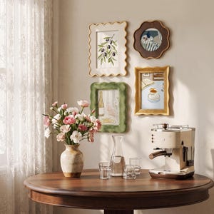 May include: A collection of four wall-mounted picture frames with wavy edges, featuring illustrations of olives, a blue vase, a bottle, and a coffee cup. Below, a ceramic vase with pink and white flowers, a glass carafe, two glasses, and a beige espresso machine.