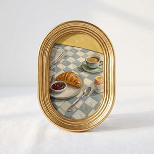 May include: An oval gold-toned frame containing a painting of a breakfast scene on a blue and white checkered tablecloth. The painting depicts a croissant, a small bowl of red jam, a cup of coffee, a soft-boiled egg in a cup, and a silver spoon.