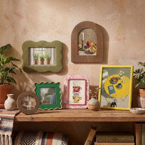 May include: A collection of various picture frames including a green wavy frame, a wooden arched frame, a small round wooden frame, a square green wavy frame, a floral frame, and a yellow beaded frame, all displayed on a wooden shelf.