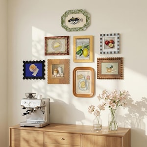May include: A collection of framed art prints with various food-themed illustrations, including lemons, cheese, and bread. The frames are in various styles and colors, arranged on a white wall. A silver espresso machine sits on a wooden cabinet.