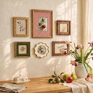 Può includere: Una parete galleria con cinque cornici decorative contenenti varie stampe botaniche, tra cui un'illustrazione centrale di una mela. Sotto, un portaburro floreale, un vaso bianco con iris viola e frutta fresca poggiano su una superficie in legno.