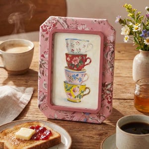 May include: A rectangular picture frame covered in pink fabric with a floral pattern, featuring an illustration of four stacked teacups in white, red, blue, and yellow with floral designs, set within a pink border on a light background.